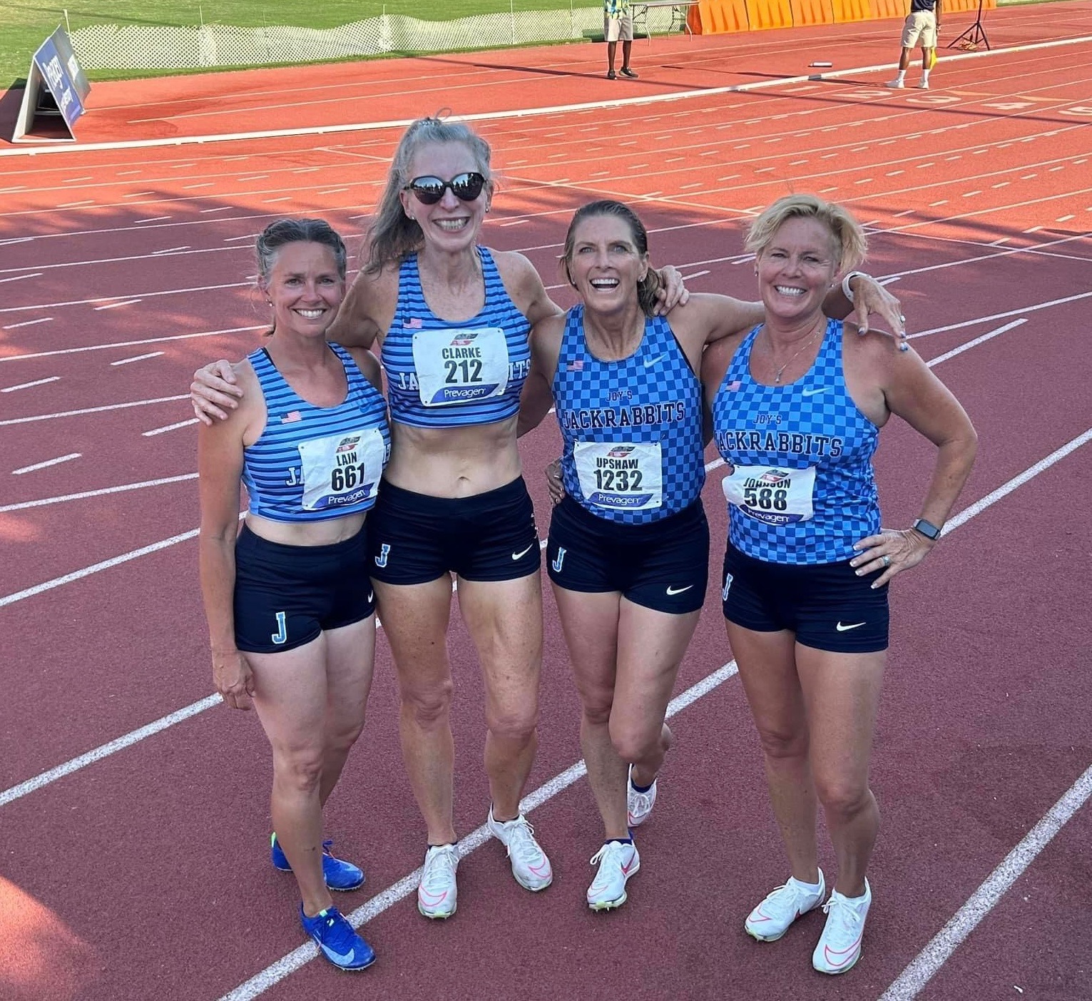 Four smiling Joys Jackrabbit Masters relay runners standing arm in arm on an outdoor track